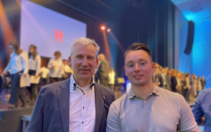 Herr Prof. Felhauer und startING-Amunus Nicolas Koessler stehen bei der Abschlussfeier zusammen vor der Bühne, im Hintergrund sieht man das Hochschul-Orchester und weitere Akteure.