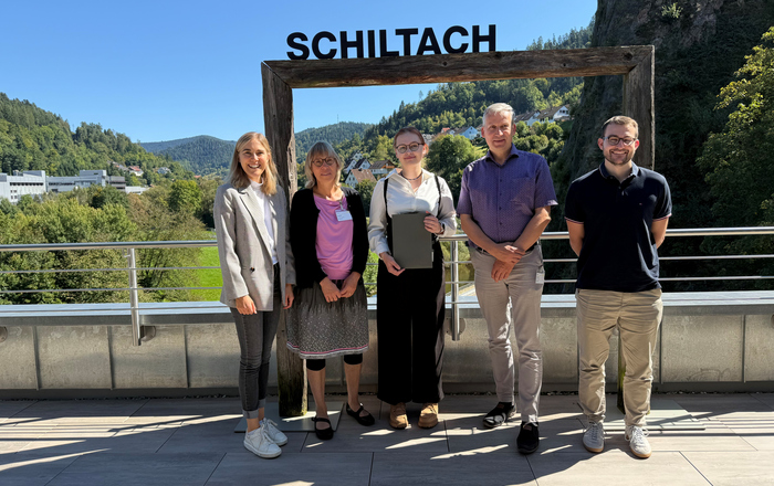 Fünf Menschen stehen bei strahlendem Sonnenschein vor einem großen Holzrahmen auf dem Schiltach steht und der eine Aussicht auf den gleichnamigen Ort einrahmt.