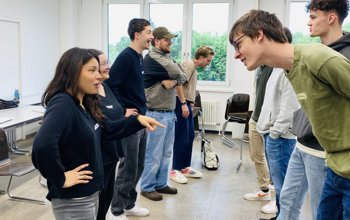 Gruppe aus Studierenden bei einem Workshop in einem Raum. In einer Reihe stehen sich immer 2 Personen gegenüber und interagieren miteinander. Im Hintergrund sind Fenster zu sehen.