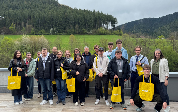 Gruppe Studierender steht mit gelben VEGA Taschen auf einer Terrasse, im Hintergrund sieht man Wald und Wiesen.