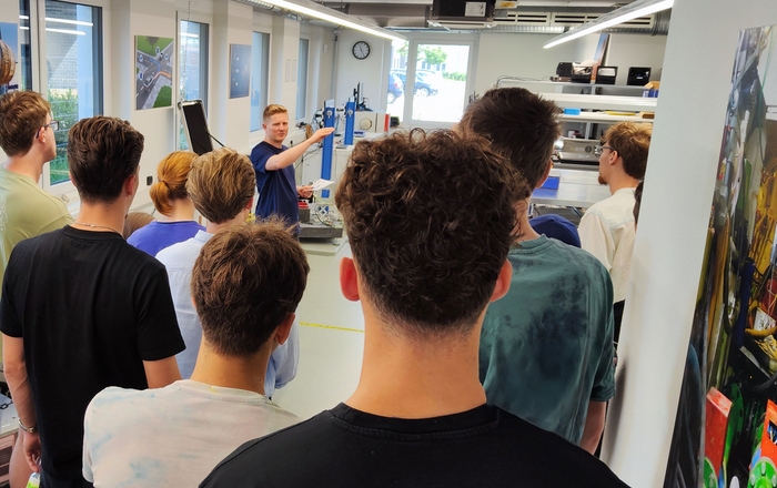 Eine Gruppe von Studierenden, die man von hinten sieht, hört dem Referent von GeneSys zu. Im Hintergrund sieht man verschiedene technische Geräte.